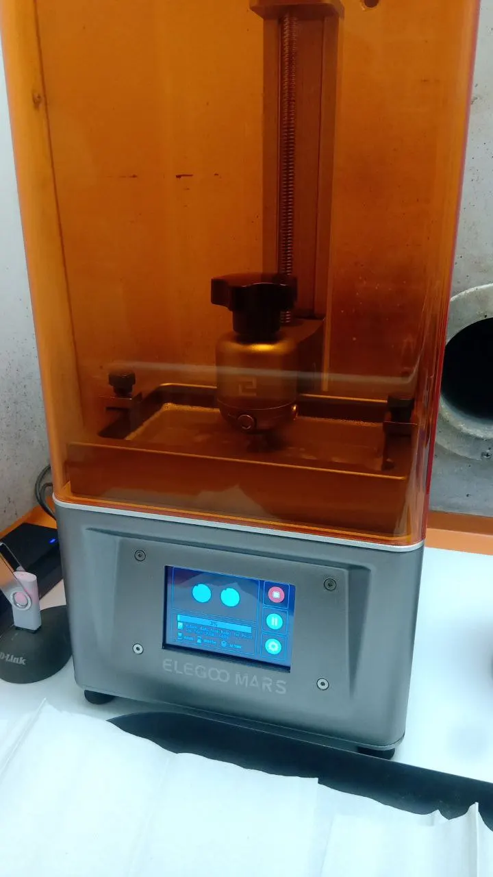 A small LCD resin printer mid-print: the part hangs from the platform above a resin tray lit from below by a screen, the same method as the Form 4