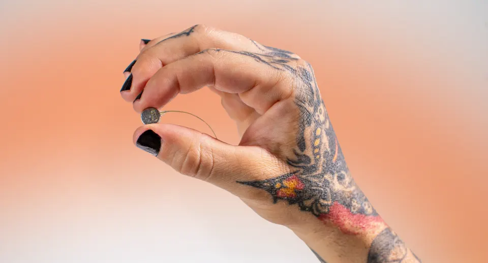 The Connexus implant held between two fingers: a coin-sized module whose tiny wire electrodes enter the brain, with a thin lead running to the chest unit