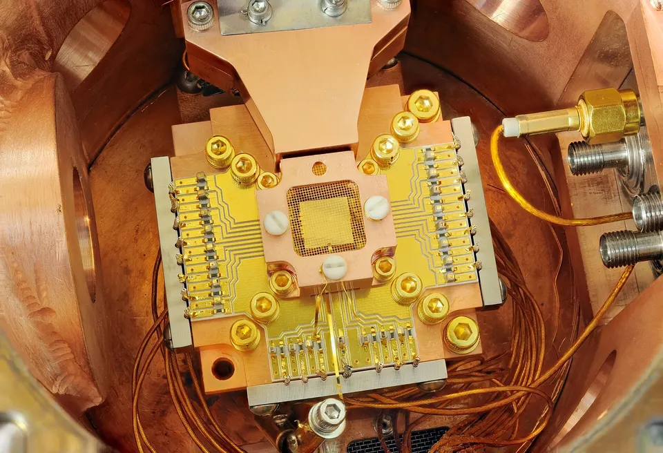 A NIST ion-trap chip in its copper mount, not Helios: charged atoms float just above the gold chip, held in place by its electrodes' voltages