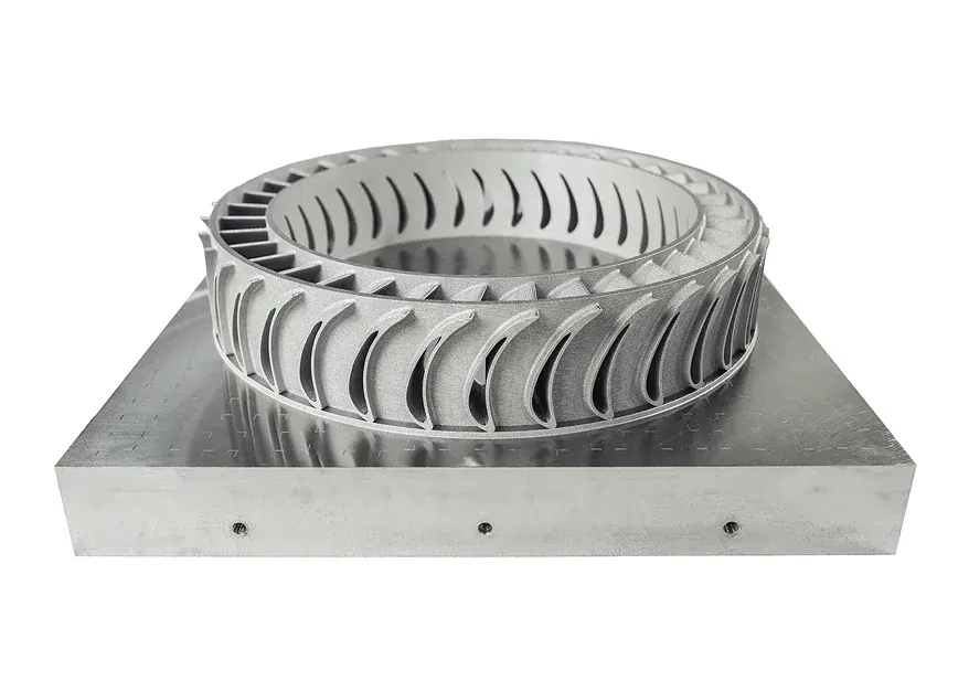 A finned metal ring still fused to its build plate: lasers melt powder layer by layer, growing the part upward from the plate